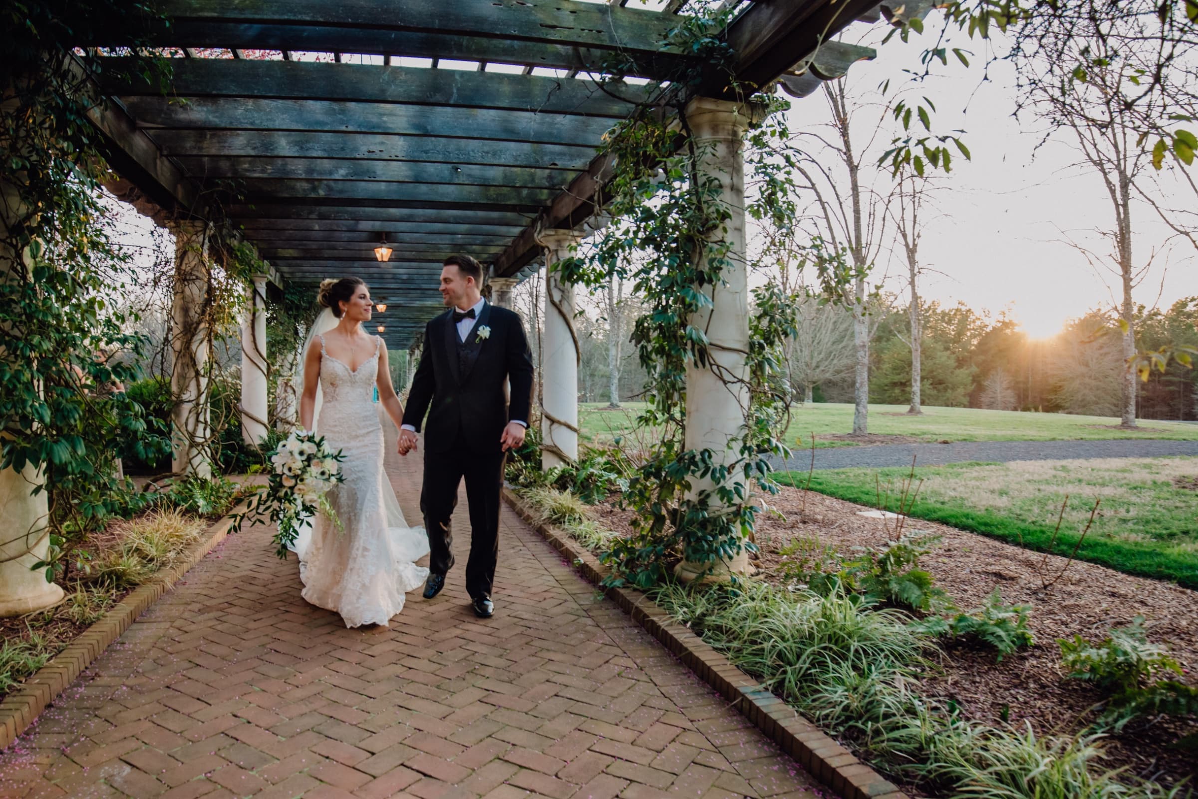 Couple under the vine-covered pergola on the brick path at sunset — Daniel Stowe Botanical Garden