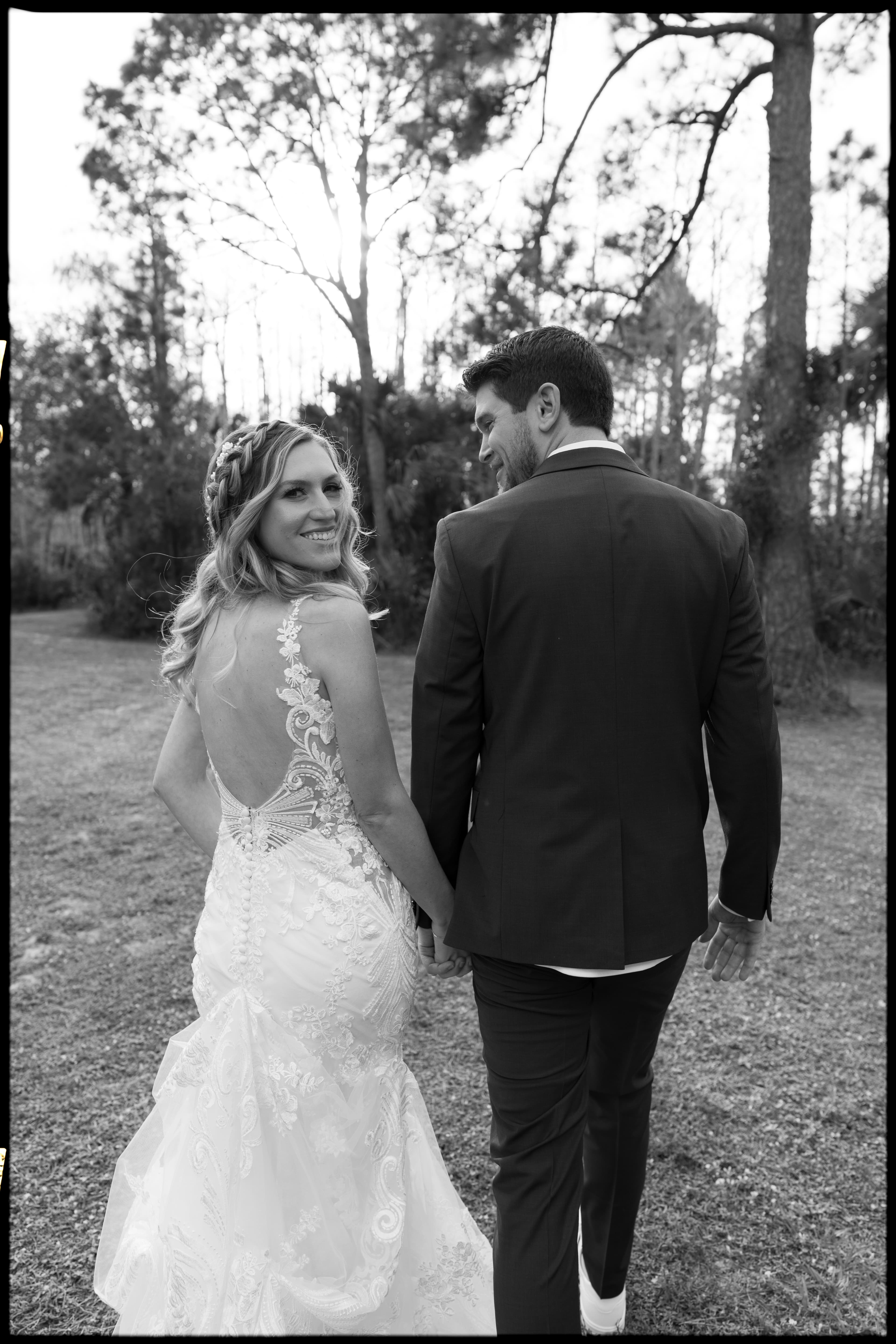 Bride glances back as she and her groom walk hand in hand through the pines — black and white Fort Myers wedding portrait