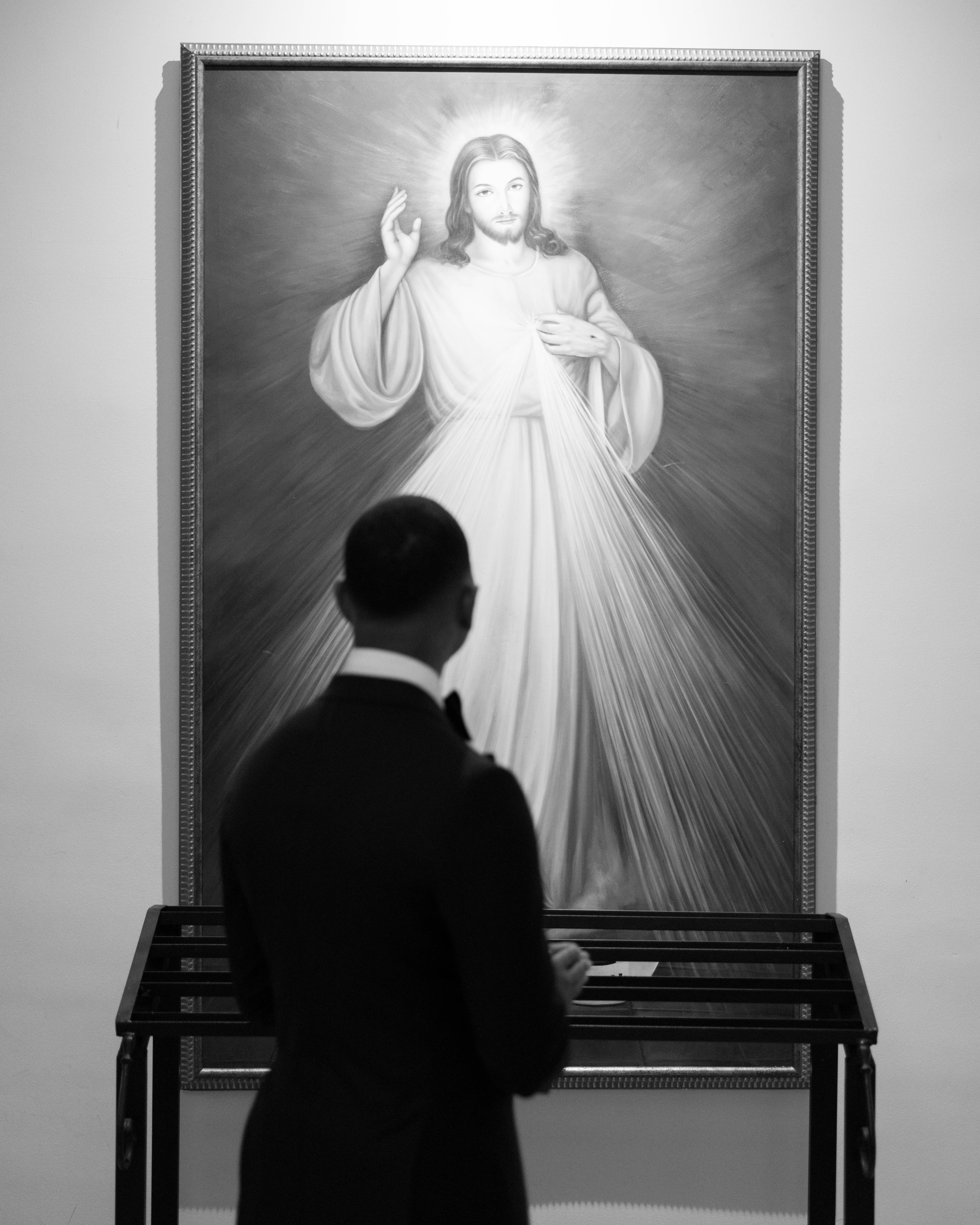 Black and white portrait of the groom pausing before a Divine Mercy painting in the church