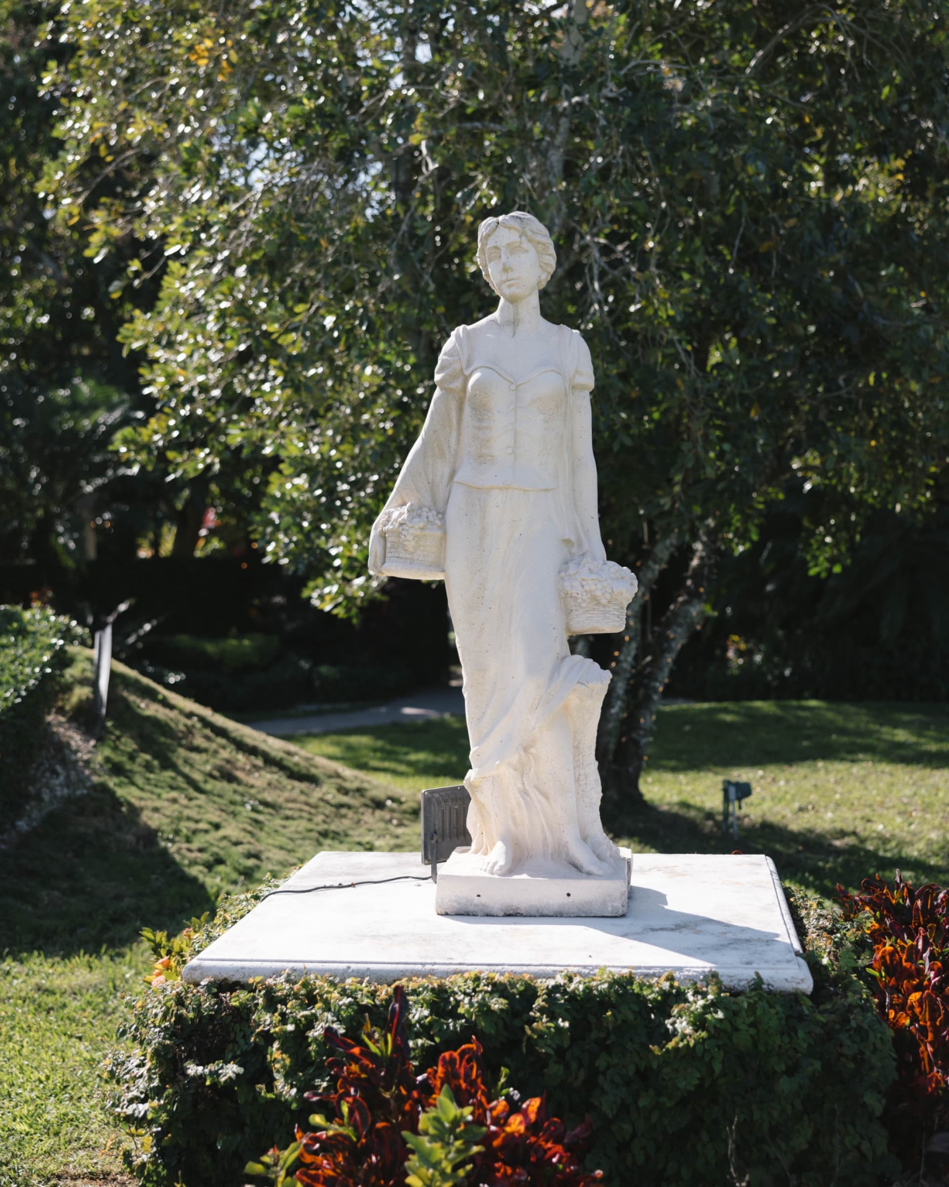 Classical garden statue on a stone plinth among clipped hedges and croton — Villa Toscana Miami