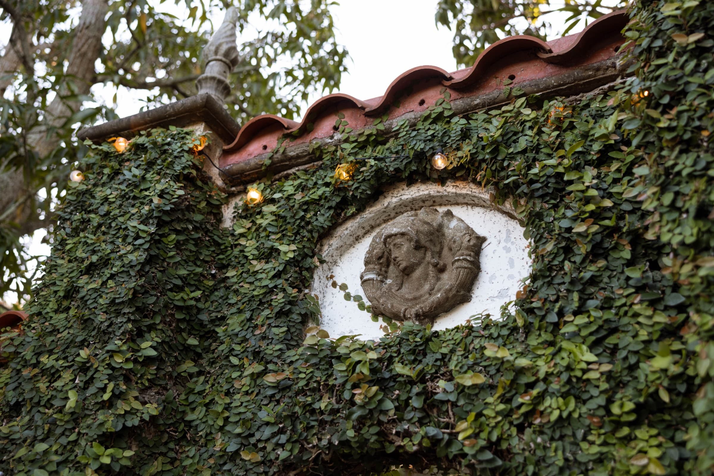 Carved stone medallion set in an ivy wall beneath terracotta roof tiles and warm festoon bulbs — Villa Toscana Miami